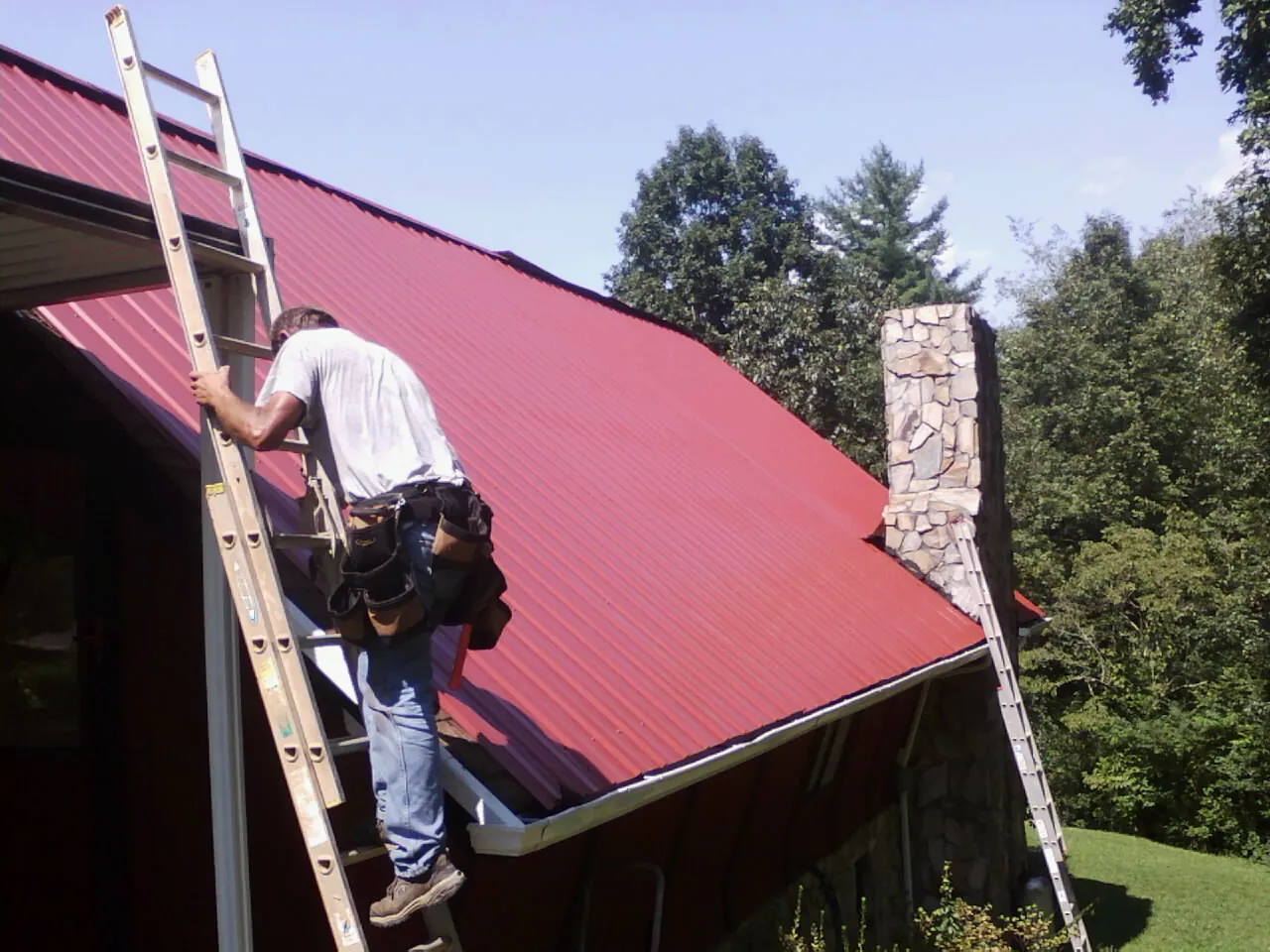 Roofer on a ladder beside a red metal roof and stone chimney — Soffit & Fascia Repair by Roofing Brothers in Fostoria